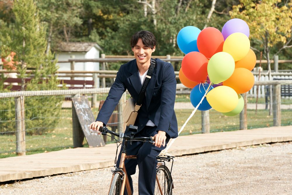 Sota Fukushi como Hiro, de terno, pedala uma bicicleta segurando balões coloridos num caminho de cascalho