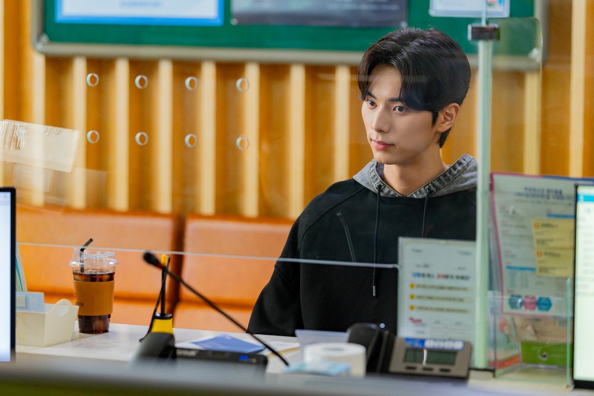 Lee Jun-ho como Kang Sang-ung sentado atrás do vidro de um balcão de atendimento, com copo de café gelado e papéis na mesa