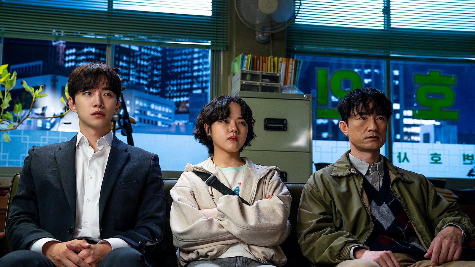 Lee Jun-ho, Kim Hyang-gi e Kim Byong-chul sentados lado a lado num escritório com janelas azuladas em cena de Cashero