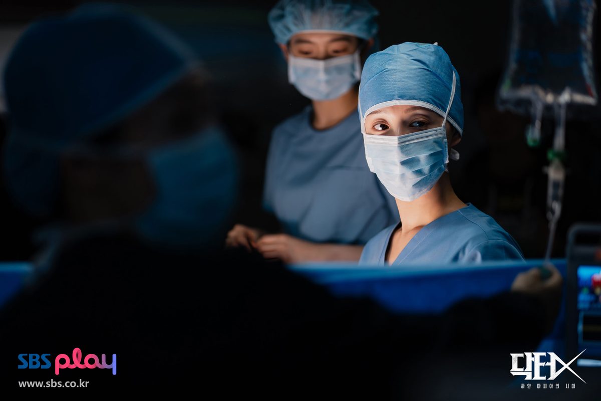 Kim Ji-won de touca e máscara cirúrgica azuis olha para a câmera durante uma operação, ao lado de outra profissional