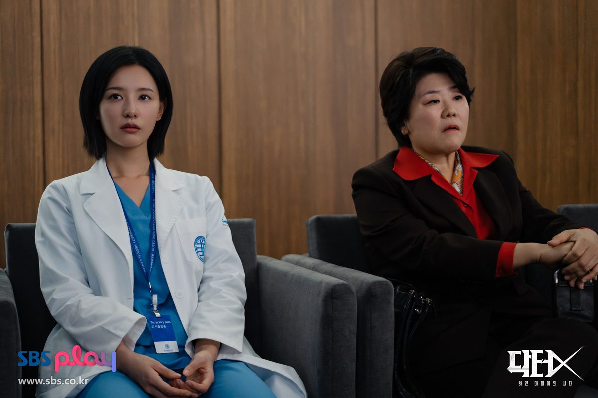 Kim Ji-won de jaleco branco e Lee Jung-eun de terno escuro e blusa vermelha sentadas lado a lado num sofá