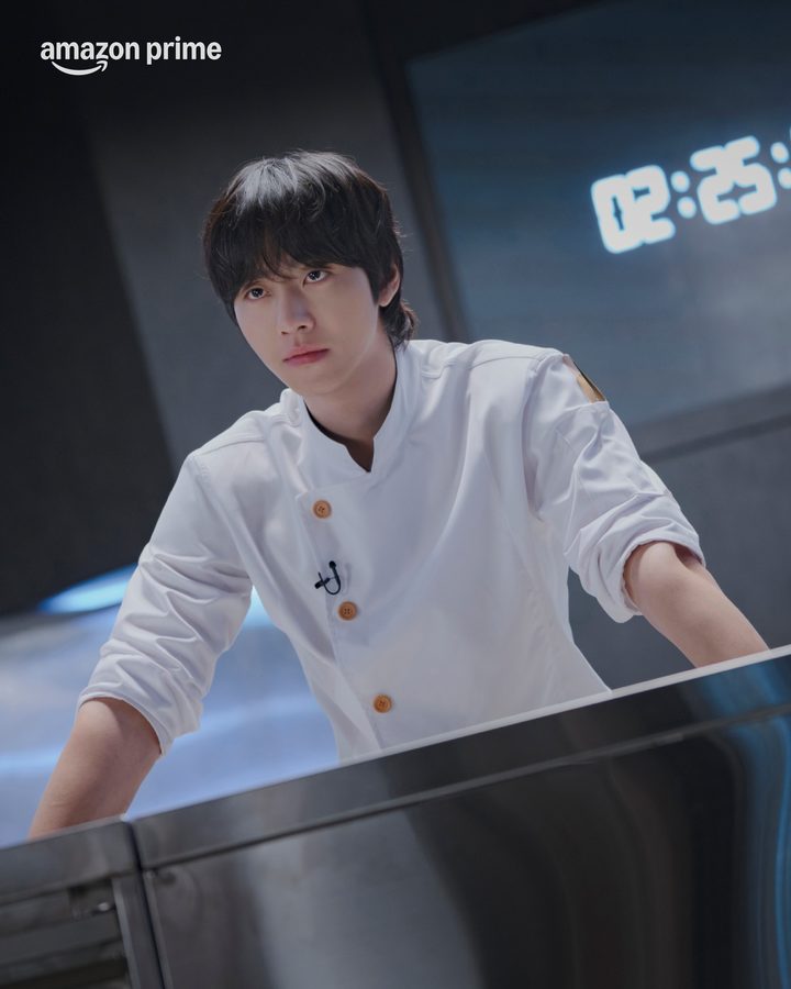Ahn Hyo-seop de dólmã branca apoiado numa bancada de aço, com um cronômetro digital ao fundo