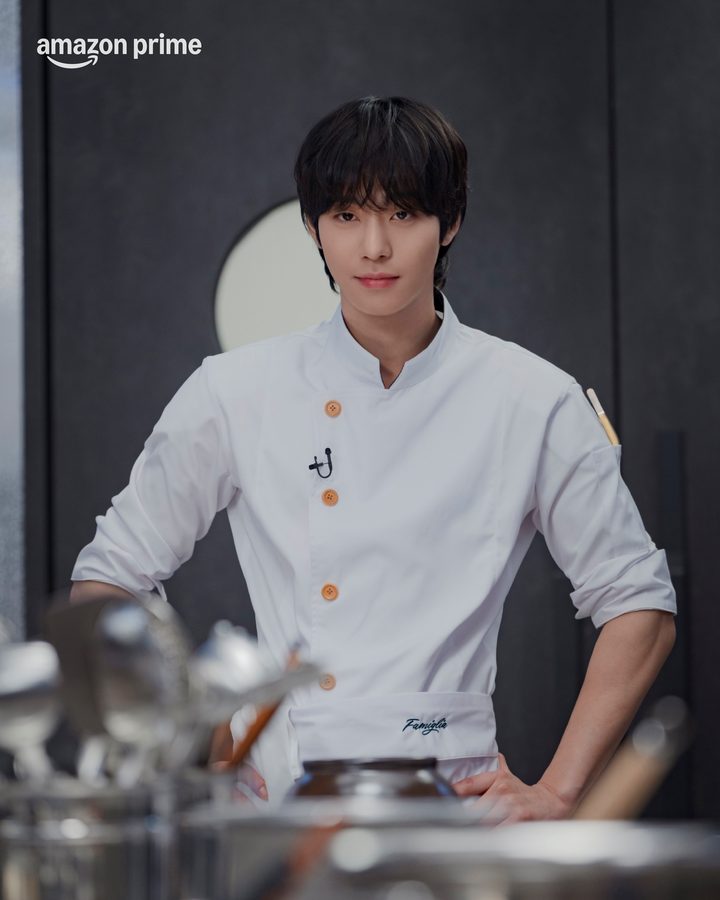 Ahn Hyo-seop de dólmã branca com botões cor de cobre, em pé numa cozinha escura, olhando para a câmera