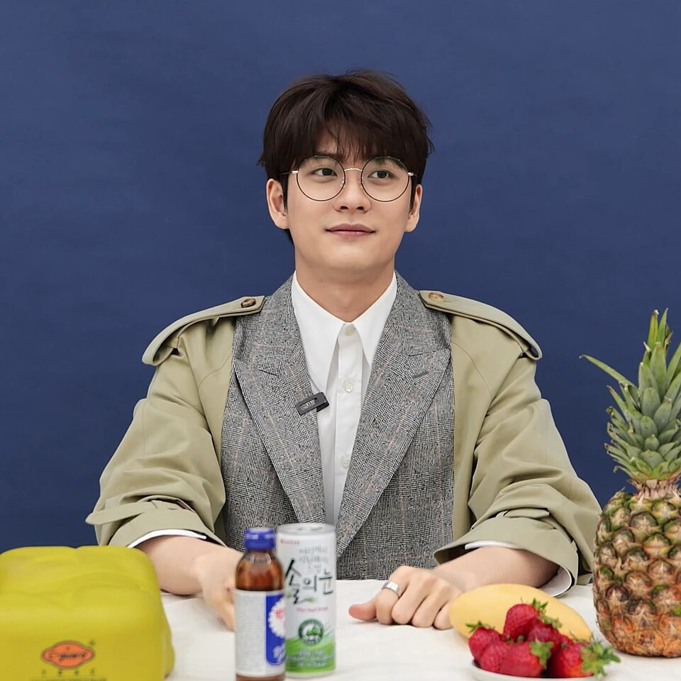 Kang Tae-oh de óculos, sentado diante de um fundo azul
