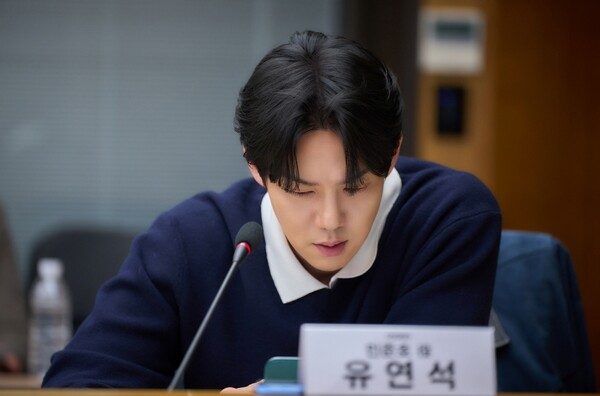Yoo Yeon-seok de suéter azul-marinho lê o roteiro de The Perfect Lie diante de um microfone e da placa com seu nome