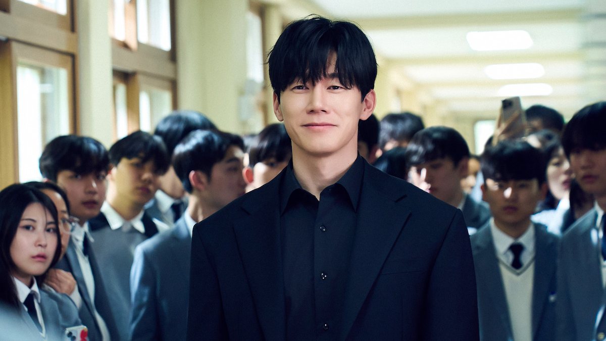 Kim Moo-yul como Na Hwa-jin, de terno preto, sorri no corredor de uma escola cercado por alunos de uniforme