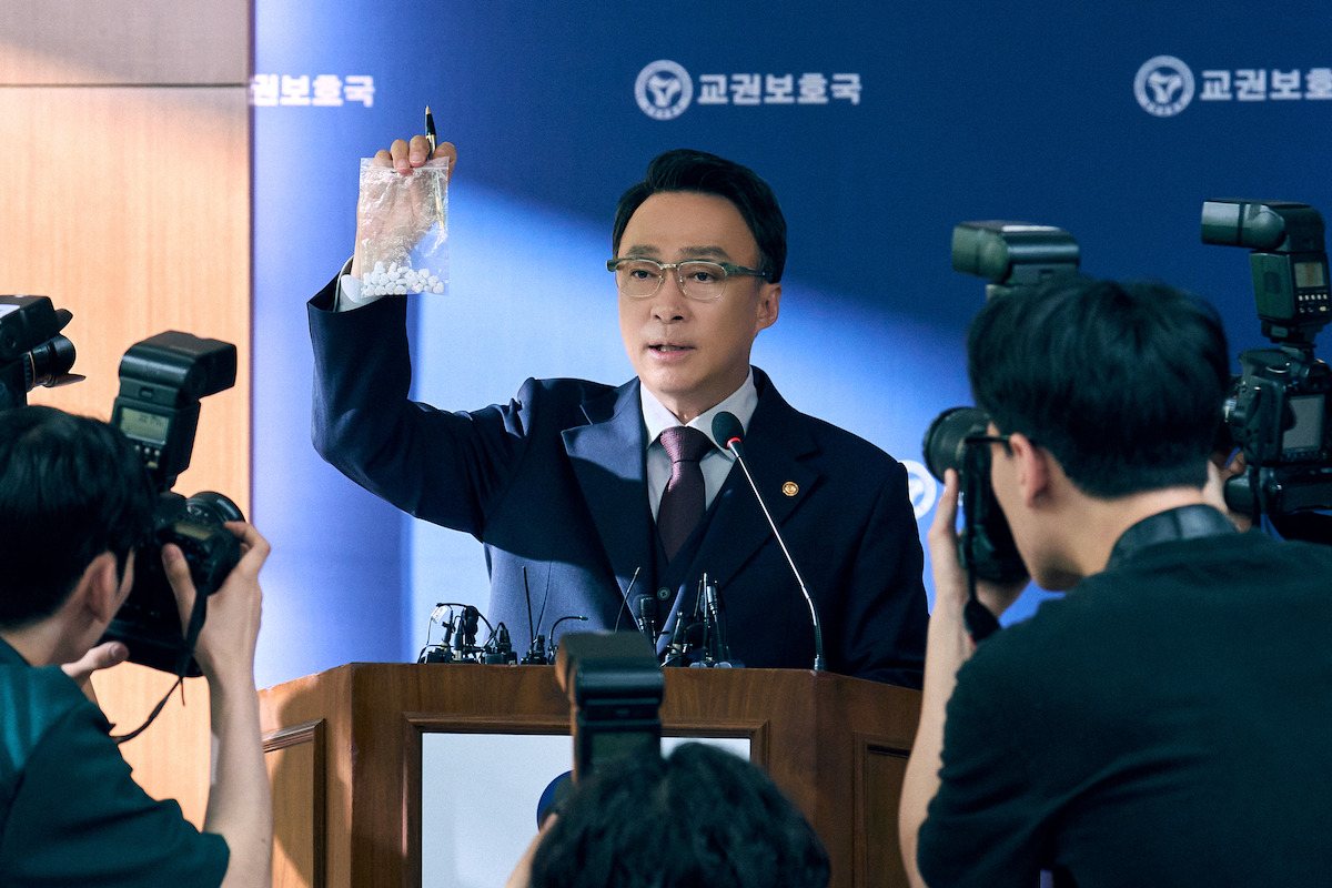 Lee Sung-min como o ministro Choi Gang-seok ergue um saco plástico diante de fotógrafos numa entrevista coletiva