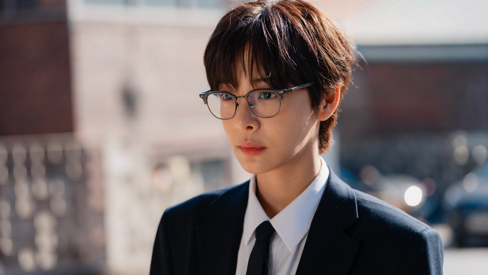 Seorina como Kang Jae-hee, de cabelo curto, óculos, terno escuro e gravata, numa rua ensolarada