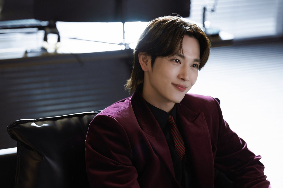Yim Si-wan como Yoon Yi-jun, sorrindo, de terno bordô e gravata listrada, sentado numa cadeira de couro
