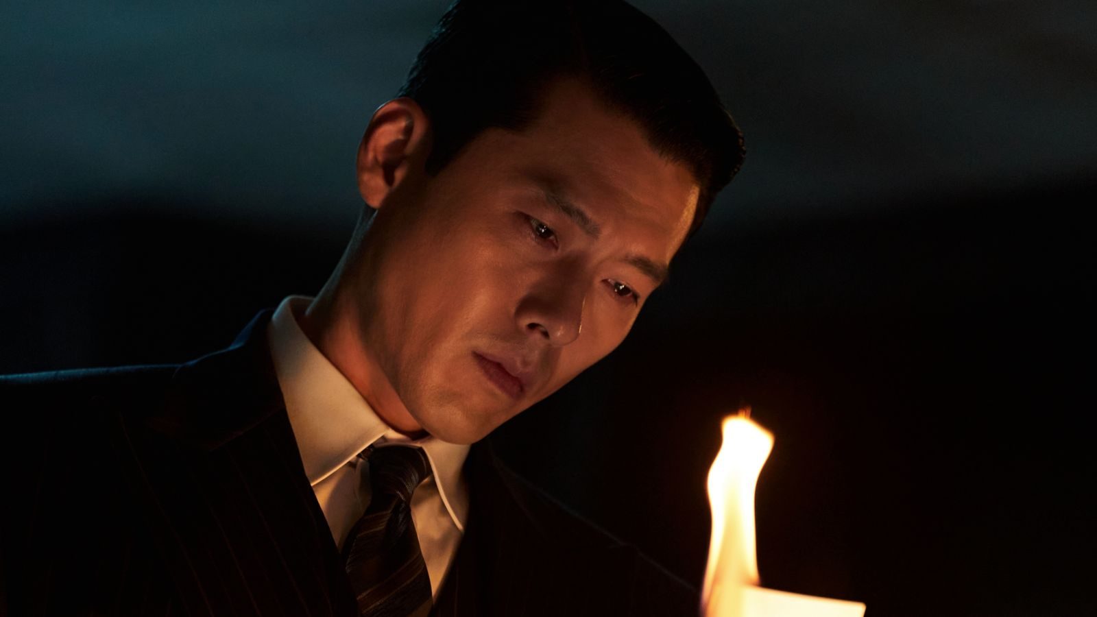 Hyun Bin como Baek Ki-tae, de terno escuro e gravata, olha para baixo diante de um papel em chamas