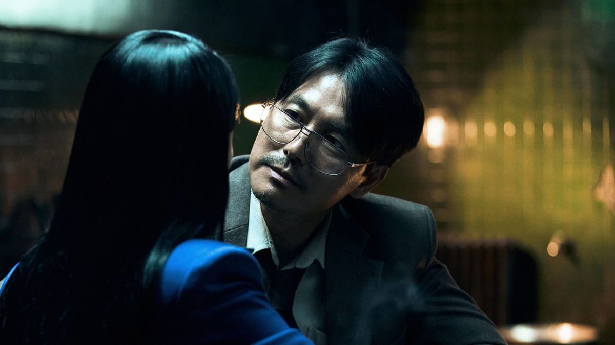 Jung Woo-sung de óculos e jaqueta azul diante de um mural com fotos e papéis presos