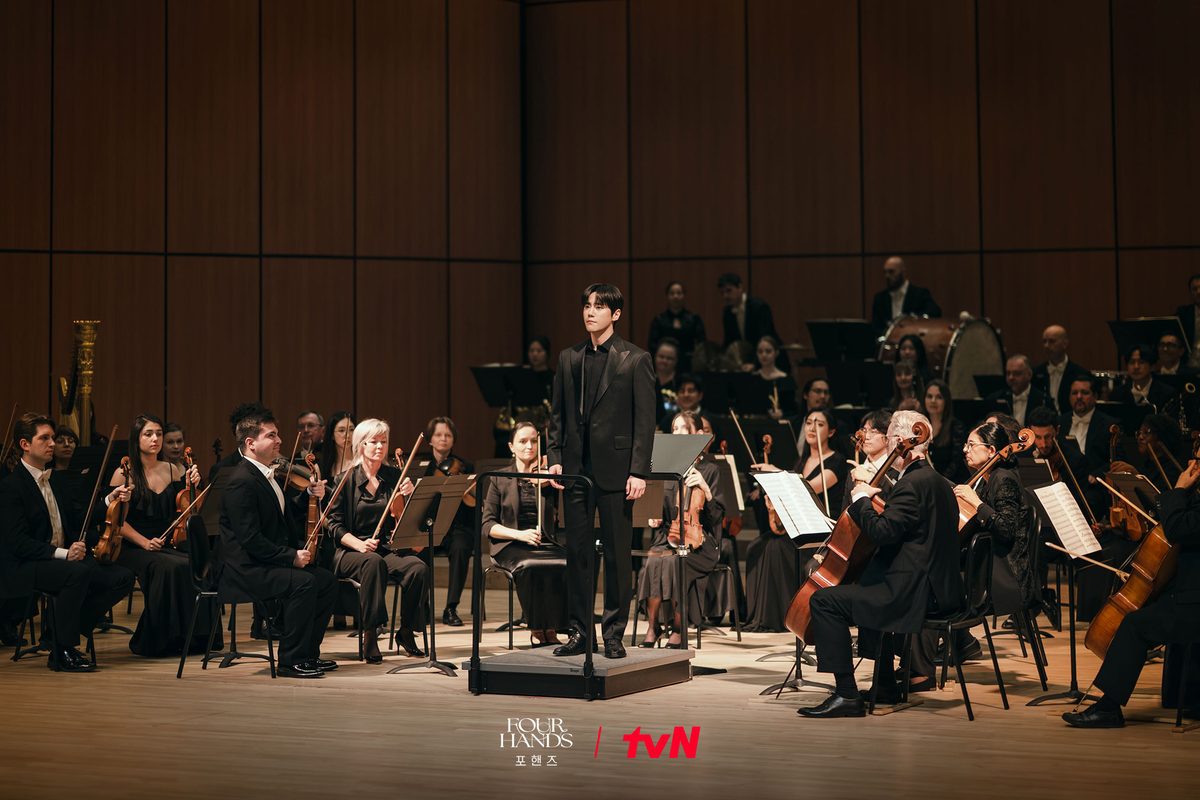 Jovem regente de terno preto à frente de uma orquestra num palco de madeira