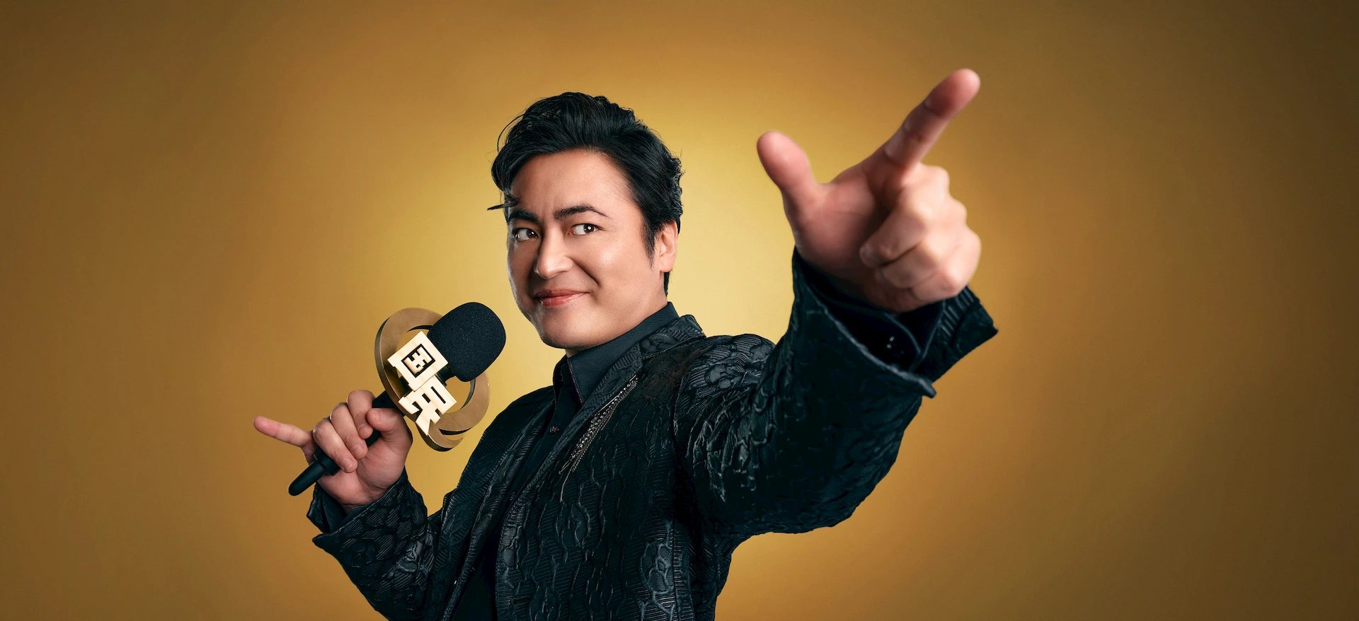 Takayuki Yamada como KK segura um microfone diante de um cenário dourado em Quiztopia
