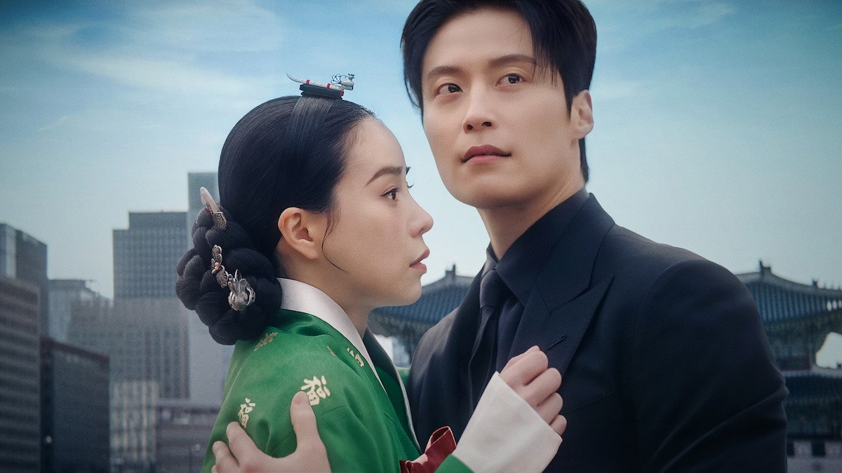 Lim Ji-yeon de hanbok verde e Heo Nam-jun de terno preto abraçados diante de prédios de Seul e telhados de palácio