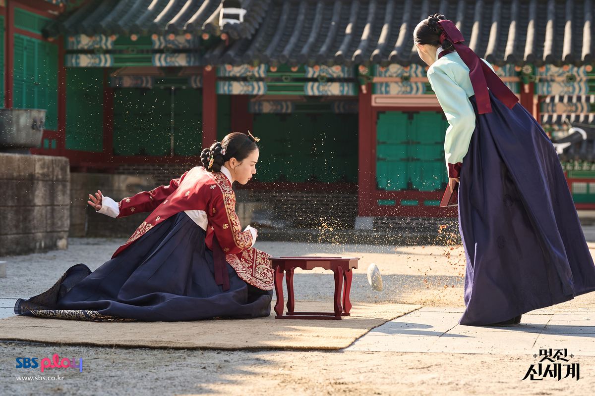 Lim Ji-yeon como Kang Dan-sim, de hanbok vermelho bordado, ajoelhada no pátio do palácio ao lado de uma mesinha diante de uma dama da corte