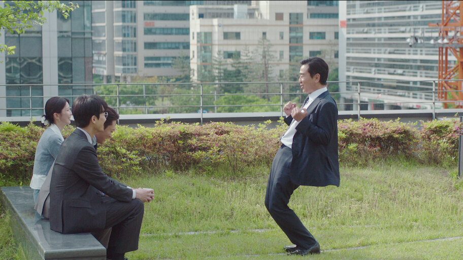 Cena de A Vida dos Sonhos do Sr. Kim em um terraço com jardim: homem de terno escuro faz pose animada diante de três colegas sentados num banco
