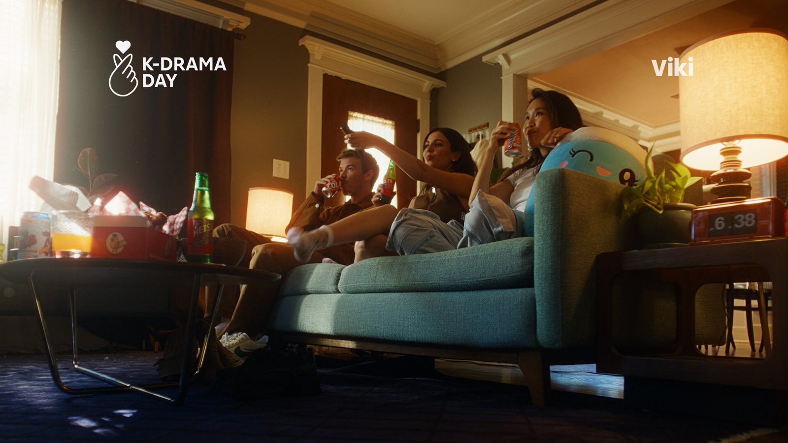 Amigos num sofá assistem à TV à noite com lanches, com os logotipos do K-Drama Day e da Viki no alto