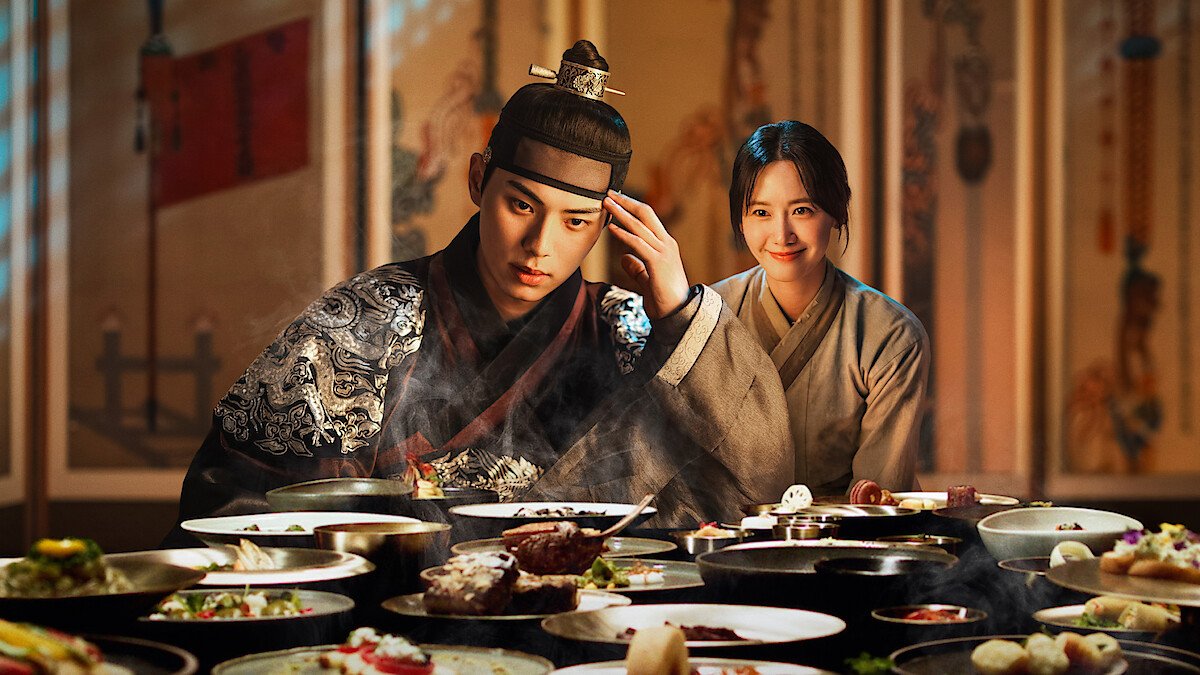 Lee Chae-min e Lim Yoon-a diante de uma mesa farta em cenário de palácio, em arte de Bon Appétit, Vossa Majestade